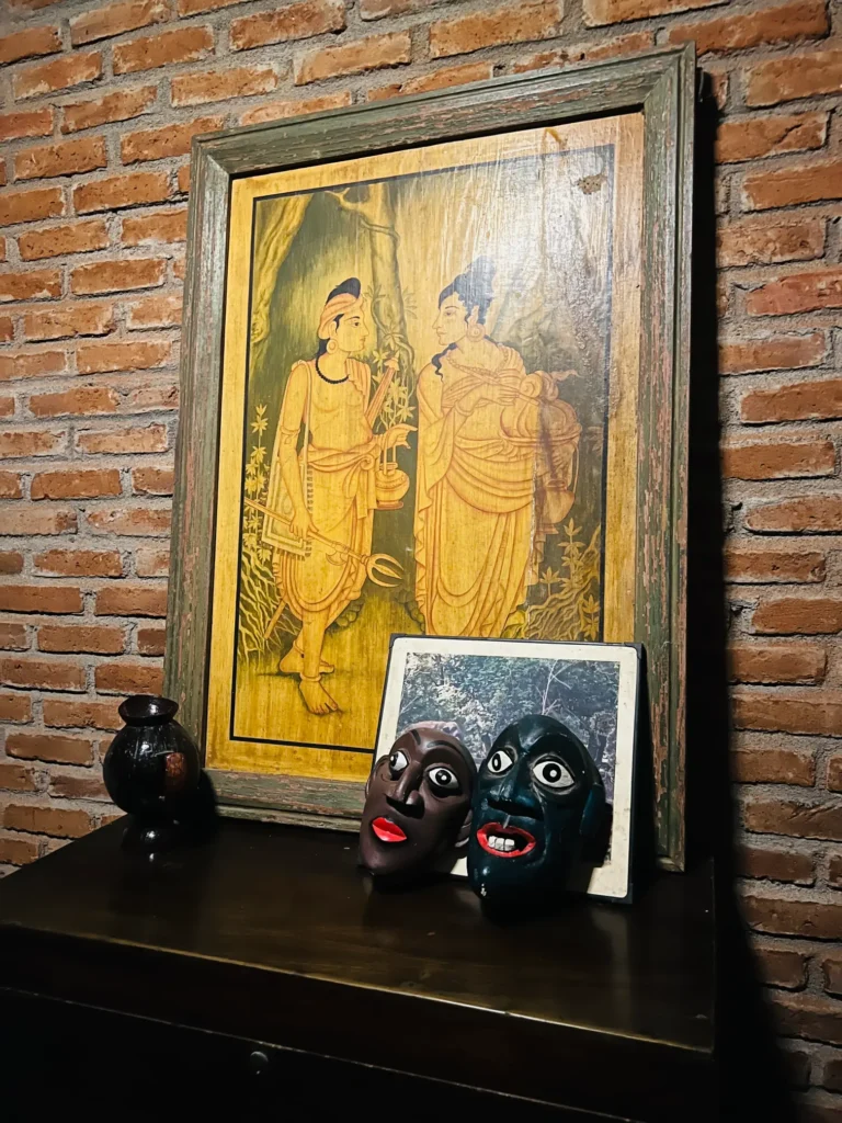 A framed painting or illustration depicting two figures in traditional Sri Lankan clothing, accompanied by two decorative masks and a small vase.